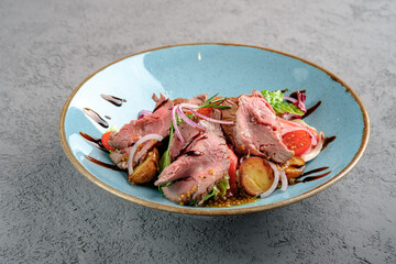 Sliced rare roast beef salad with roasted baby potatoes, fresh greens, red onion rings, cherry tomatoes and balsamic glaze in light blue bowl. Gourmet beef salad