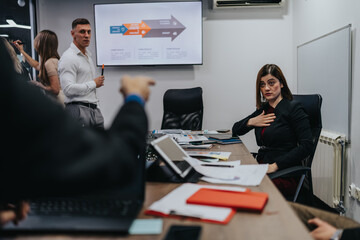 A man presents with a pointer while a woman sits at a conference desk. Colleagues work on documents and devices in a bright, contemporary meeting room.