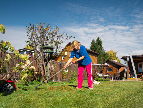 Woman raking cut grass in a lush green backyard garden near a wooden house. Summer outdoor work and home improvement concept.