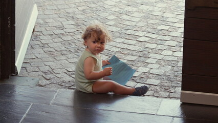 Curious toddler seated near open door looking back while holding paper, facing camera with thoughtful expression as cobblestone street extends beyond threshold