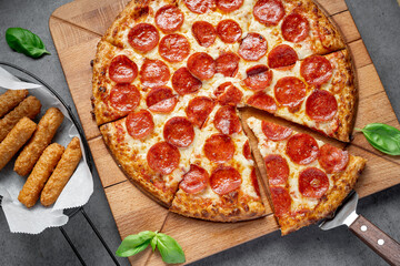Slices of pepperoni pizza on wooden board with mozzarella sticks nearby. A wooden board holds pepperoni pizza with one slice cut out and mozzarella sticks on the side.