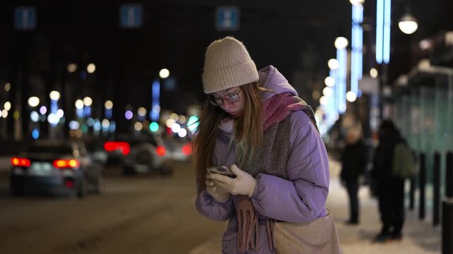 Young woman in warm winter clothes waiting for public transport at a bus stop on a cold, snowy night, using her smartphone and looking for the bus with traffic lights bokeh in the background