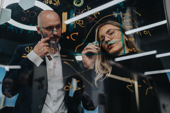 Two professionals analyze complex equations on a glass board during a collaborative session. The scene conveys focus, teamwork, and problem solving in a modern office.