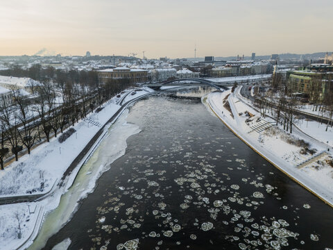 Aerial view of the Neris River cutting through a snow-dusted city, with ice floes drifting downstream under a pale winter sky, Vilnius, Vilnius County, Lithuania.