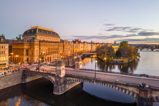 Aerial view of the National Theatre in Prague during the evening. The theatre is lit up, and the surrounding embankment and bridges are visible.