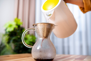 Person pours hot water from white kettle into glass Chemex with metal filter. Barista prepares fresh arabica drink at home. Morning ritual, lifestyle and energy concept