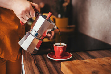 Italian moka pot pours hot espresso into red ceramic cup in kitchen. Young woman in orange clothes...