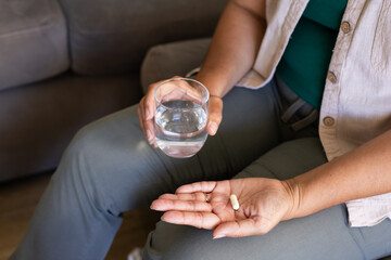 Palm holding pale capsule and metal ring while opposite hand holding water glass on gray sofa