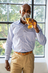 Senior African American man sipping from tall glass of orange-juice at multi-pane window in shirt