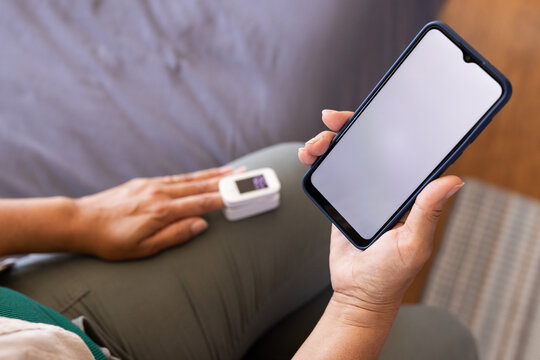 Mature adult woman sitting on bed wearing green top olive pants holding phone and pulse oximeter