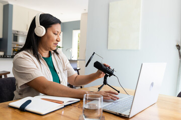 Mature adult woman adjusting microphone while working on laptop at kitchen table wearing headphones
