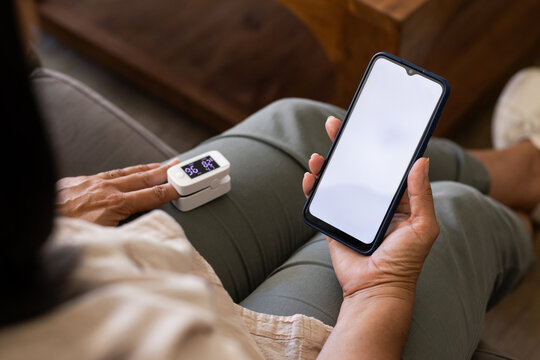 Hands holding smartphone and fingertip pulse oximeter resting on lap on sofa, soft lighting