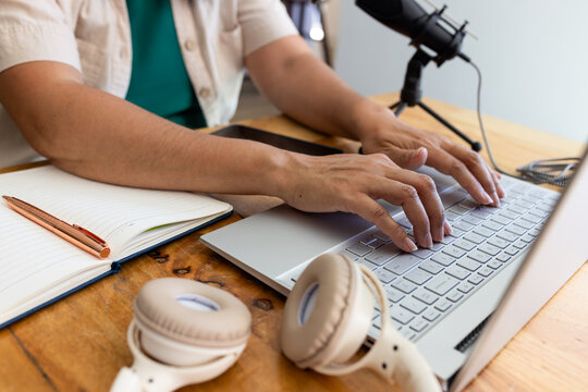 Silver laptop is sitting on wooden desk with lined notebook, copper pen, phone, headphones, mic