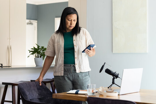 Mature Asian woman standing, holding phone, resting left hand on chair at dining table with mic