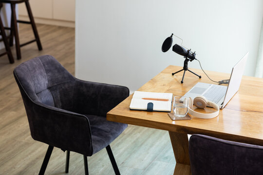Wooden desk is hosting home studio setup with laptop mic on tripod, headphones, notebook, glass