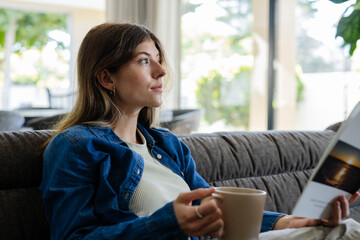 Woman sitting on tufted sofa at home in denim shirt holding beige mug, booklet, gazing outside
