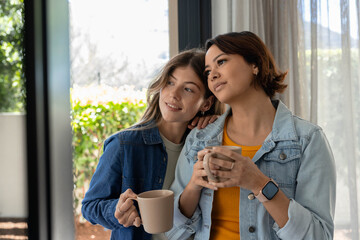 Female partners in 20s and 30s standing at glass door wearing denim jackets, holding ribbed mugs