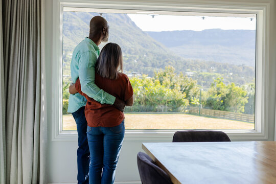 Mature couple standing at large picture window in dining room, holding each other near table