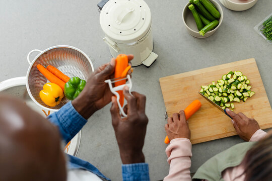 Diverse couple preparing vegetables on grey counter, peeling carrots with Y-peeler on wooden board