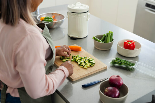 Zucchini being sliced on wooden cutting board on gray countertop, with knife and compost canister