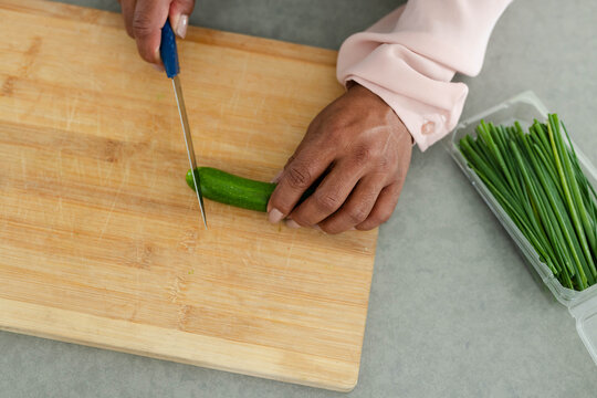 Paring knife is slicing small zucchini on cutting board on gray counter, clamshell chives nearby