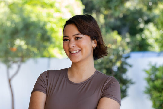 African female standing in garden with white fence, smiling, wearing brown top and hoop earrings