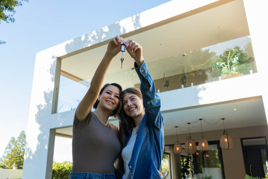 Diverse female friends standing at front of modern two-story house, holding keyring aloft