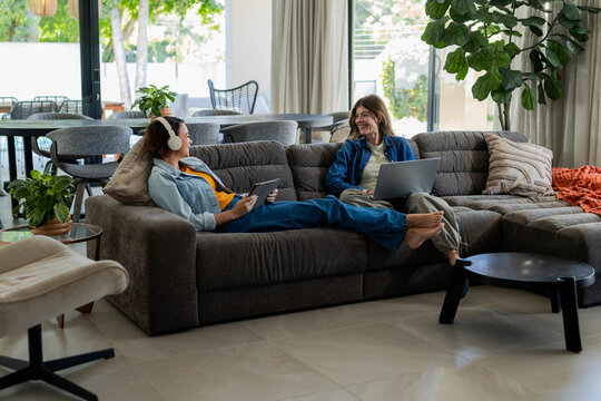 Diverse female friends relaxing on gray sofa, wearing headphones and using laptop, tablet