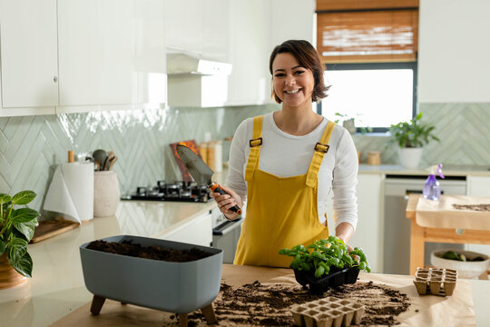 African American woman in mustard overalls potting seedlings at kitchen counter with metal trowel