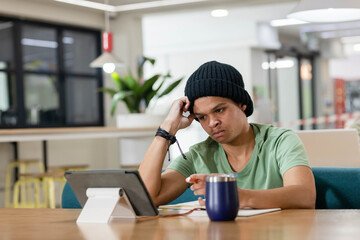 African American man in black beanie resting head, focusing on tablet at cafe table with notebook
