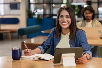 Diverse coworkers working in communal workspace, woman writing in notebook using tablet stand