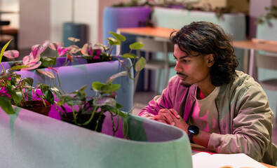 Indian man leaning forward, clasping hands by papers at lounge table with planter, copy space
