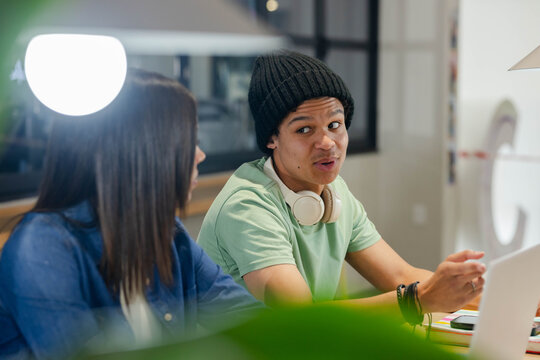 Coworkers sitting at shared table in study area, talking with laptop, headphones, desk lamp