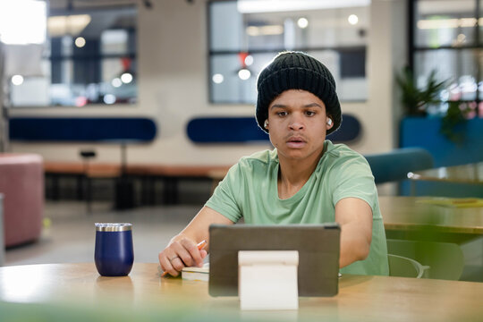 African American man studying and writing on tablet at communal workspace with notebook pen tumbler