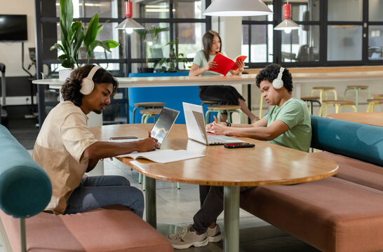 Two African American men studying at communal table, study lounge, using laptops, white headphones