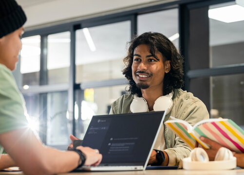 Male peers collaborating while smiling and using laptop with white headphones in study space