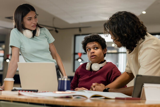 African American peers discussing around laptop at coworking table using notebook and headphones