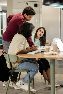 Diverse students leaning over round wooden table pointing to open book and laptop with coffee cup