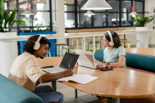 African American peers wearing headsets, sitting at coworking table, writing and typing on laptops