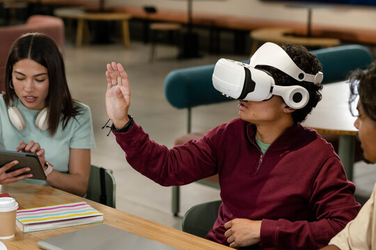 African American students sitting in campus lounge around table using VR headset, tablet, laptop