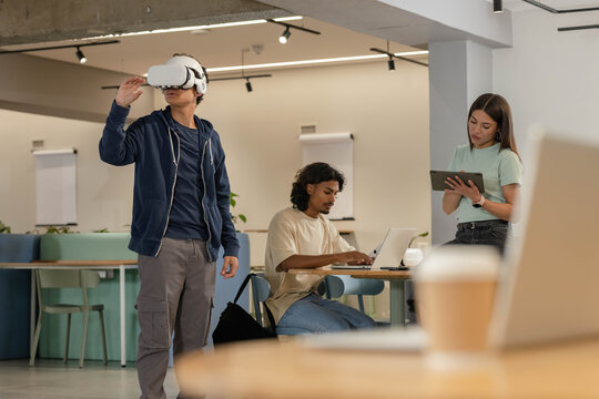 Three colleagues collaborating in co-working lounge, using VR headset, laptop, tablet, copy space