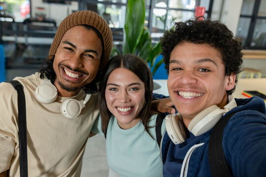 Student friends posing for selfie in campus common room with white headphones and backpacks