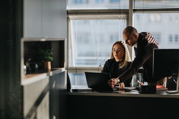 Two professionals work together at a desk, one guiding the laptop. A modern office scene with blinds, monitors, and documents.