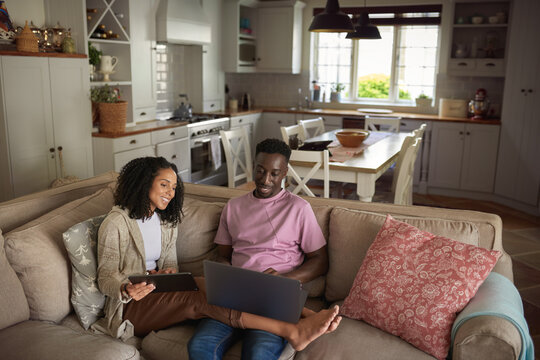 Smiling young multiracial couple using a laptop and tablet on their sofa
