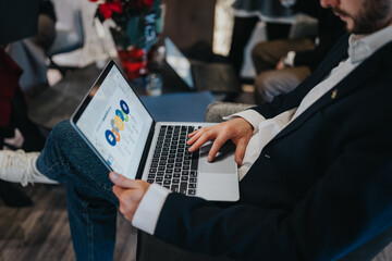 A man in a blazer uses a laptop displaying colorful charts. He sits in a relaxed, modern lounge during a casual business moment.