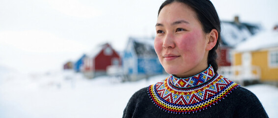 Authentic portrait of a young Greenlandic woman wearing a traditional beaded collar in a snowy Arctic village with colorful houses.