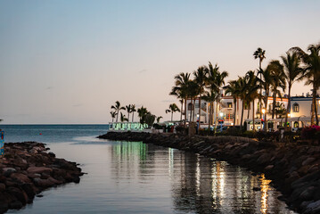 Dusk at Puerto de Mogan, Gran Canaria shows a narrow inlet to the Atlantic, rocky breakwaters, tall palms, and whitewashed buildings with warm lights and long reflections. © Aerial Film Studio