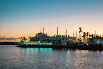 Fototapeta premium White harbor building with turquoise lights reflects on calm water. Palm trees, sailboat masts, breakwater, and people on terraces in a Canary Islands marina.