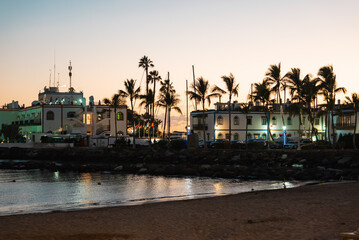 Whitewashed buildings with blue trim line Puerto de Mogan harbor on Gran Canaria. Palm trees and a sandy beach frame calm water as warm lights reflect at dusk. © Aerial Film Studio