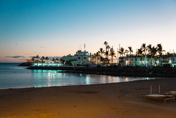 Twilight shows a small harbor with palm trees, white buildings, and a lighthouse like pier in the Canary Islands, lights reflecting on calm water and sandy beach. © Aerial Film Studio